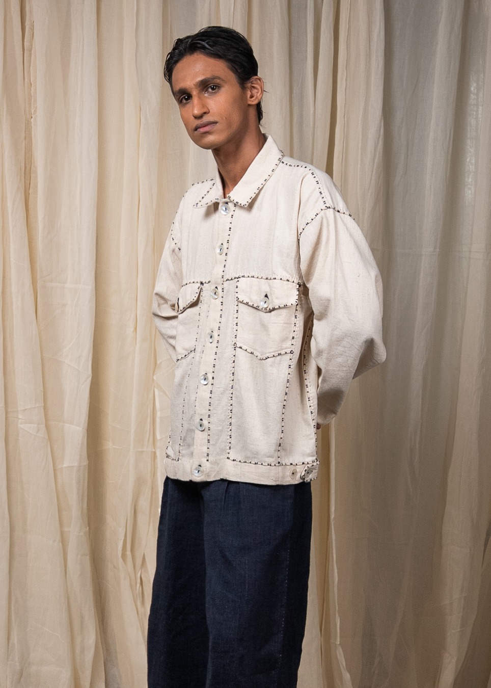 A man wearing a cream-colored linen jacket with decorative brown stitching and dark navy trousers, standing in front of a draped beige fabric backdrop.