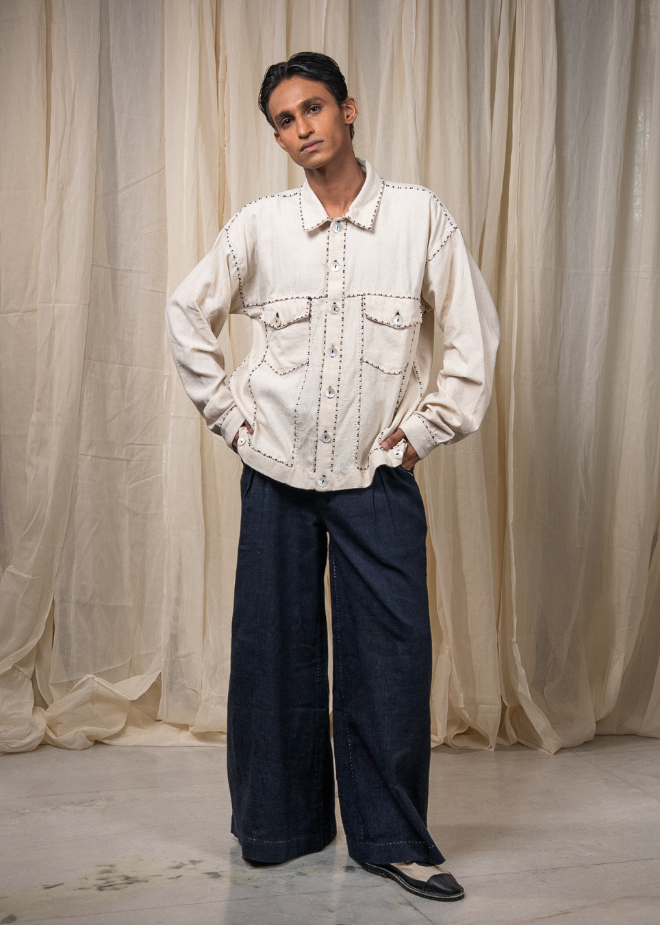 A model stands against a draped beige fabric backdrop wearing a cream-colored, long-sleeve denim jacket with decorative stitching and dark wide-leg denim pants.