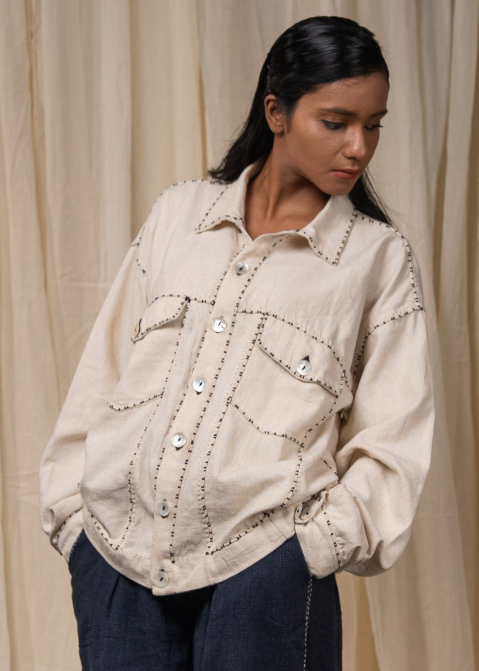 A woman wearing a cream-colored linen button-up shirt jacket with decorative dark brown stitching and silver buttons, paired with dark blue jeans, standing in front of a beige curtain backdrop.