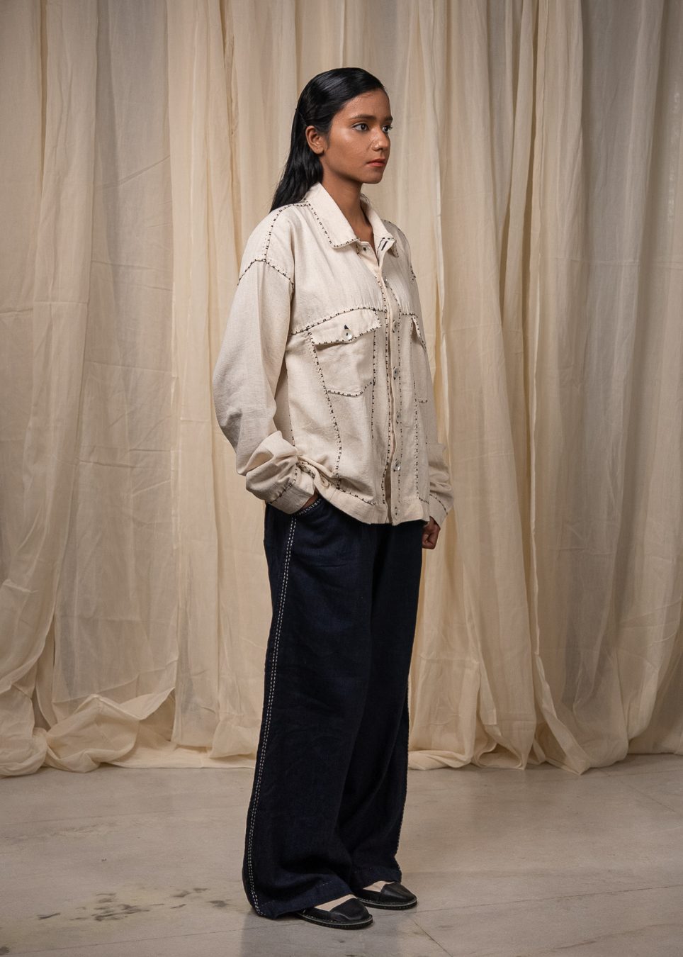 A model stands in profile wearing a cream-colored embroidered button-up shirt and dark navy wide-leg trousers, posed against a backdrop of draped beige fabric.