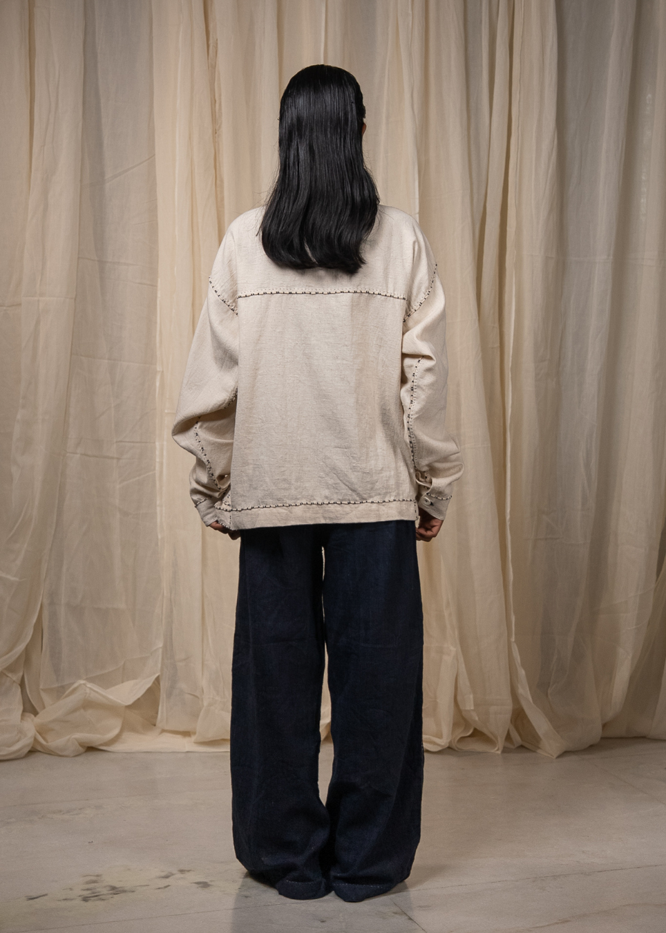 A person standing with their back to the camera, wearing a light beige oversized jacket and dark navy wide-leg pants, set against a backdrop of draped beige fabric curtains.