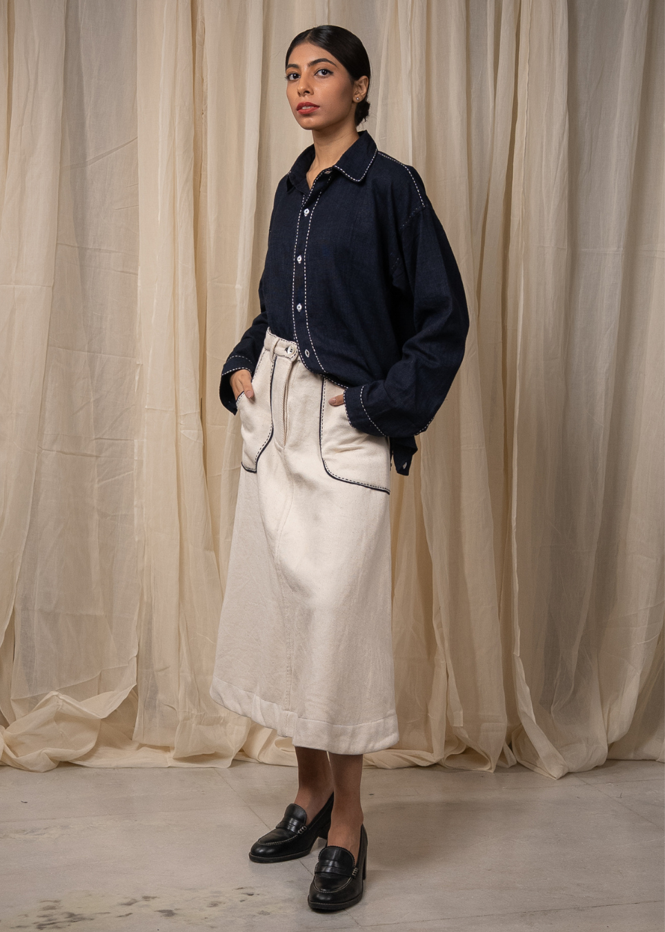 A model stands in front of draped beige fabric, wearing a dark navy blue denim shirt tucked into a cream-colored linen midi skirt with dark contrast stitching, paired with black leather loafers.