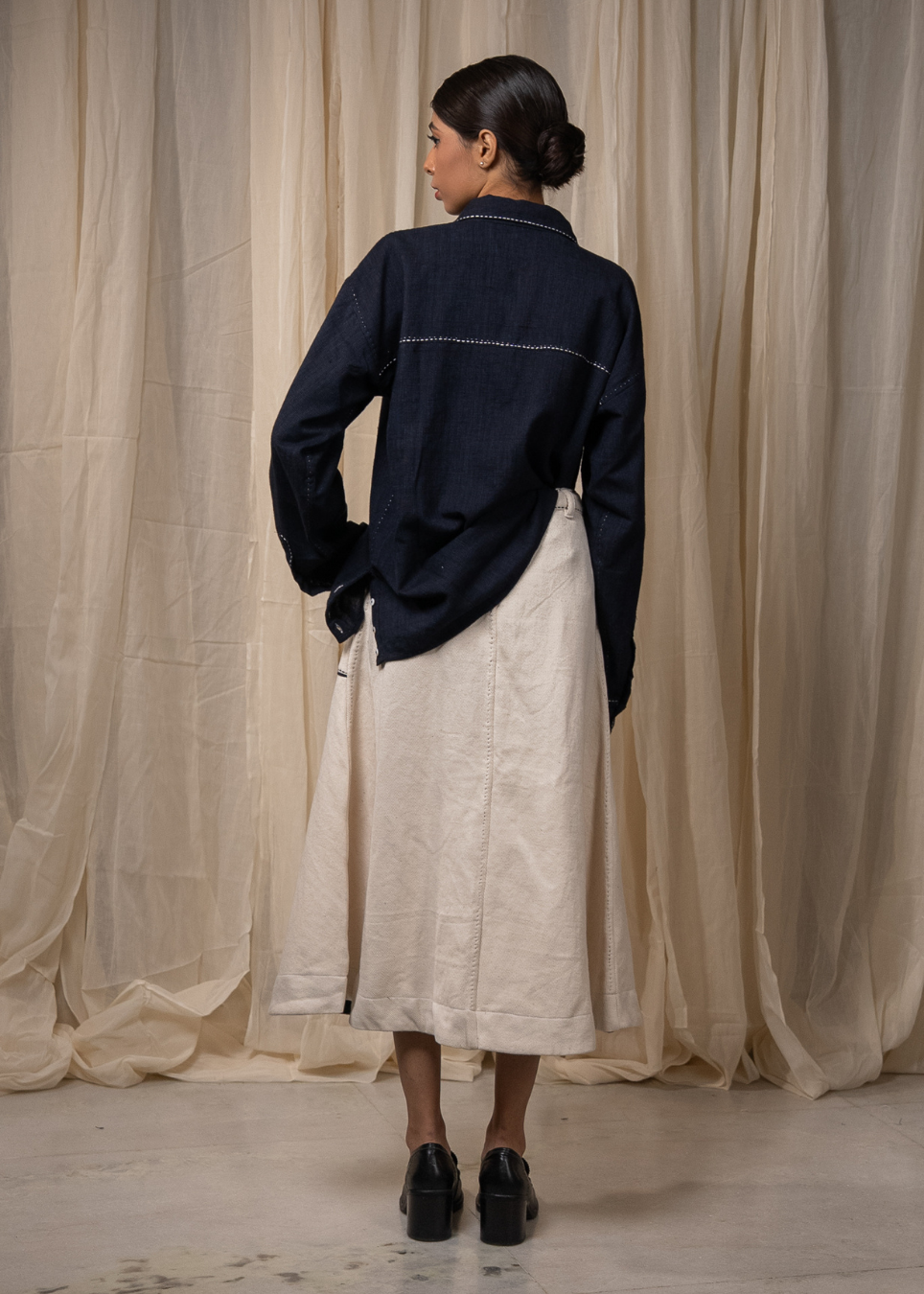 A woman stands with her back to the camera wearing a dark navy blue long-sleeve shirt with contrast stitching and a light beige linen midi skirt, posed in front of draped beige fabric curtains.