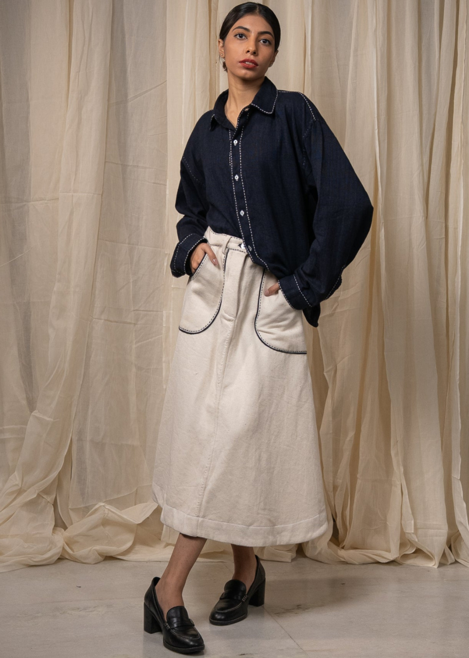 A woman models a dark navy blue long-sleeve button-up shirt with white contrast piping, paired with a cream-colored linen midi skirt and black loafers, standing in front of draped beige fabric.