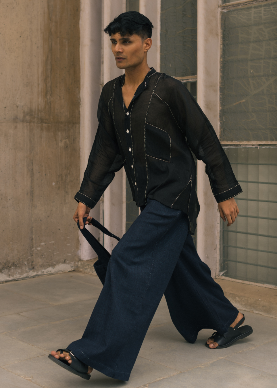 A man walking outdoors wearing a black long-sleeve button-up shirt with contrast stitching and a large chest pocket, paired with loose dark blue wide-leg pants and black sandals.