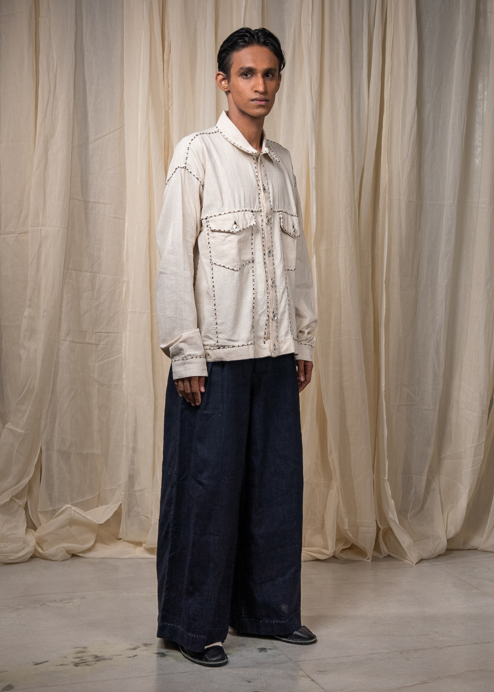 A model stands in front of draped beige fabric, wearing a cream-colored embroidered jacket with dark wide-leg trousers and black shoes.