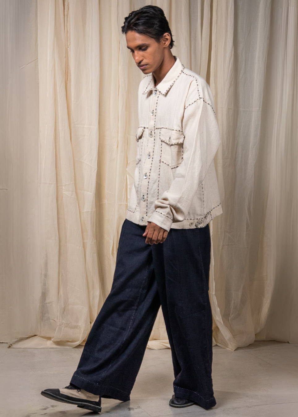 A model wearing a cream-colored embroidered jacket and dark wide-leg jeans, standing in front of a draped beige fabric backdrop.