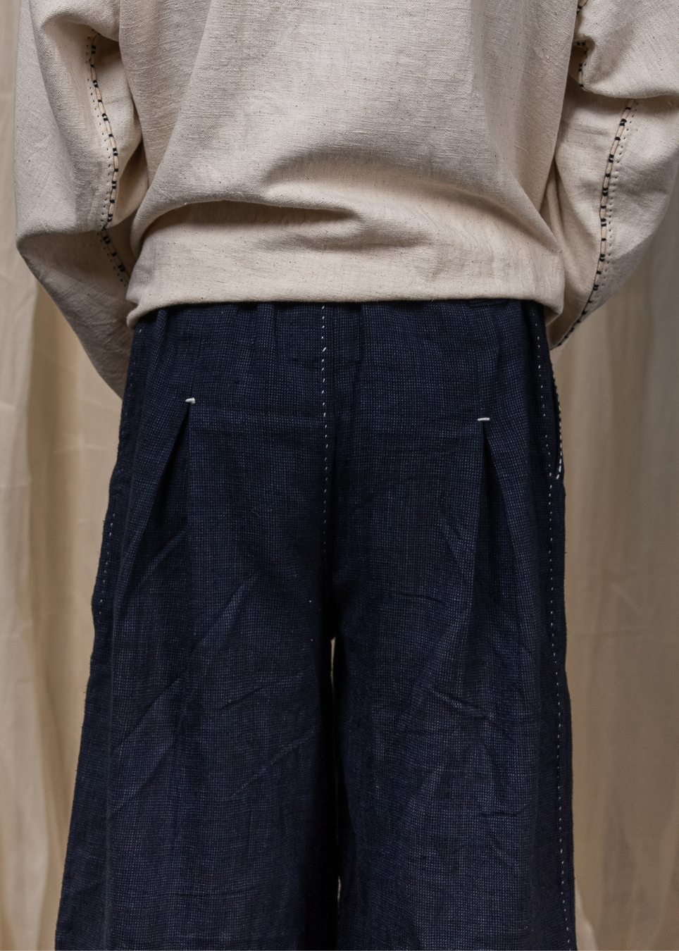 Close-up back view of a person wearing dark navy wide-leg pleated trousers and a light beige long-sleeve top, standing against a neutral draped fabric backdrop.