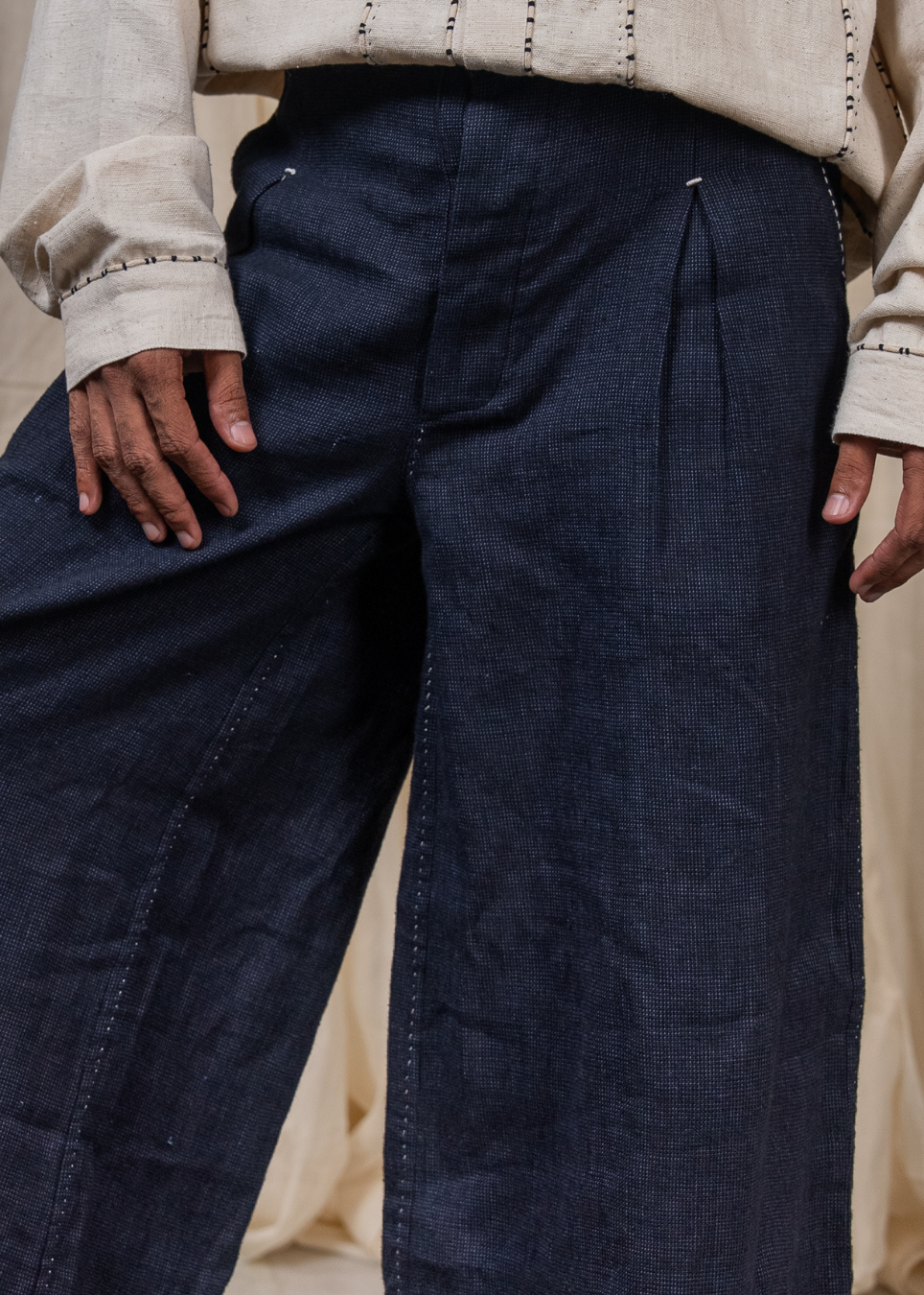 Close-up of a person wearing dark indigo wide-leg trousers and a cream embroidered shirt, with hands resting on the thighs.