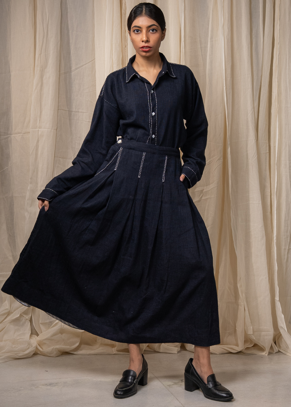 A person wearing a dark indigo denim long-sleeve shirt and matching wide-leg skirt with white contrast stitching, standing against a beige fabric backdrop.