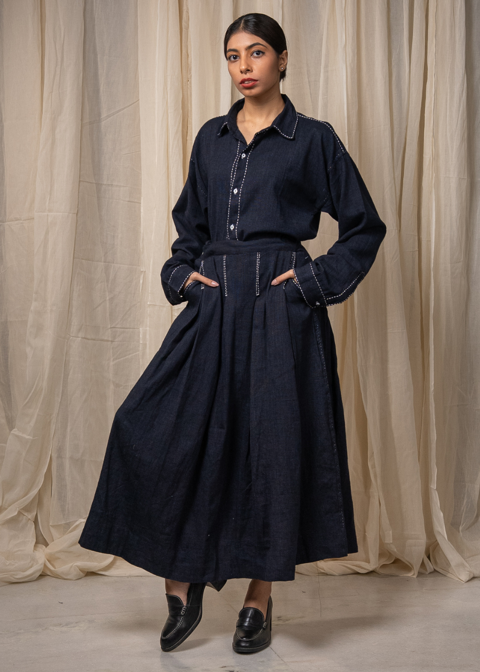 A person wearing a dark indigo denim long-sleeve shirt and matching wide-leg skirt with white contrast stitching, standing against a beige fabric backdrop.