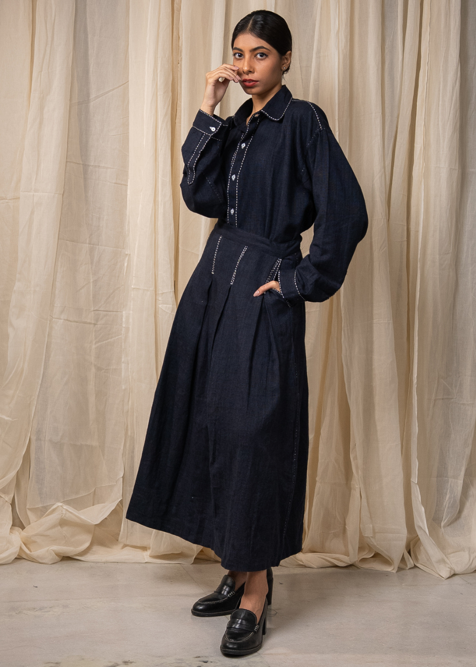 A person wearing a dark indigo denim long-sleeve shirt and matching wide-leg skirt with white contrast stitching, standing against a beige fabric backdrop.