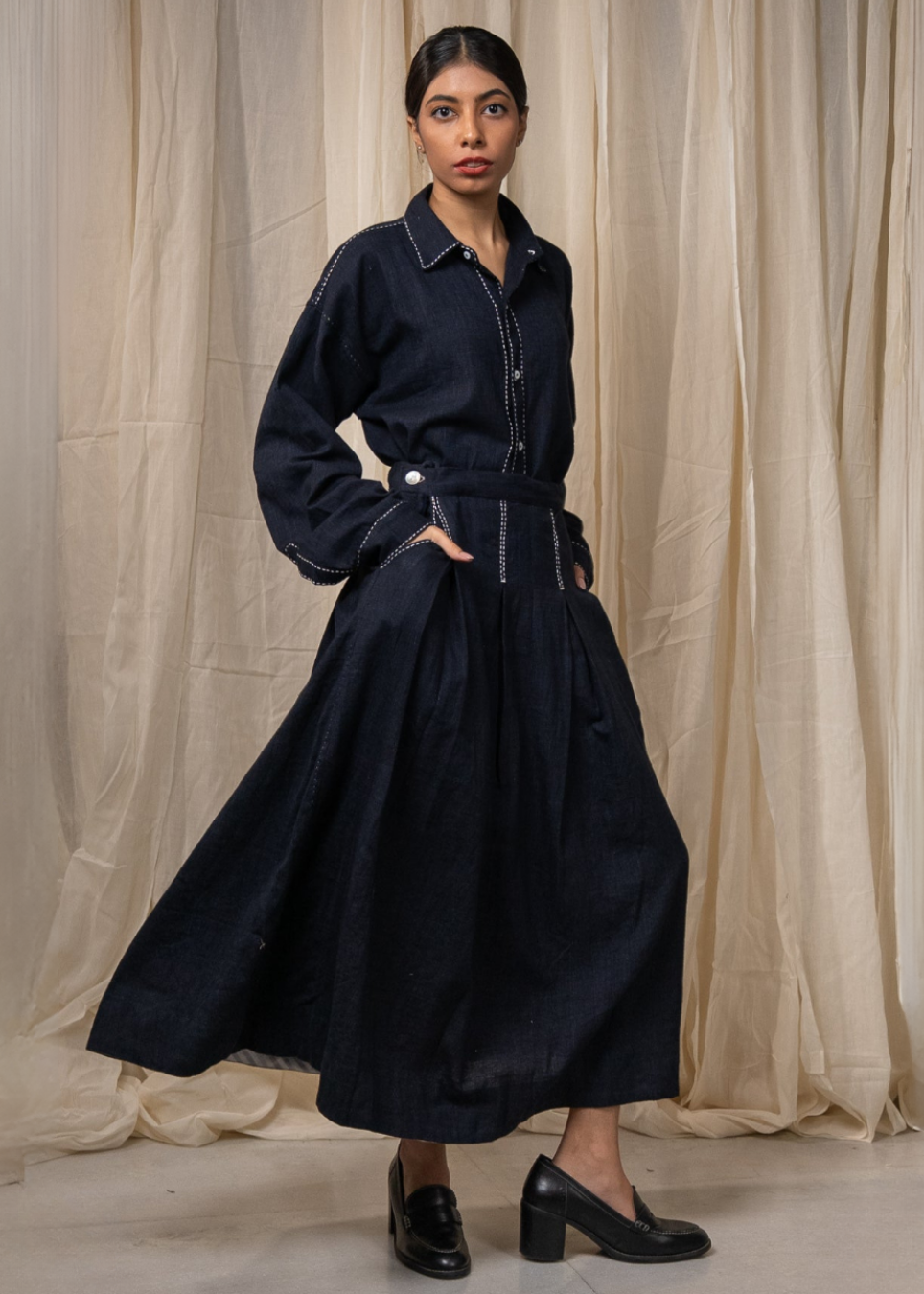 A person wearing a dark indigo denim long-sleeve shirt and matching wide-leg skirt with white contrast stitching, standing against a beige fabric backdrop.