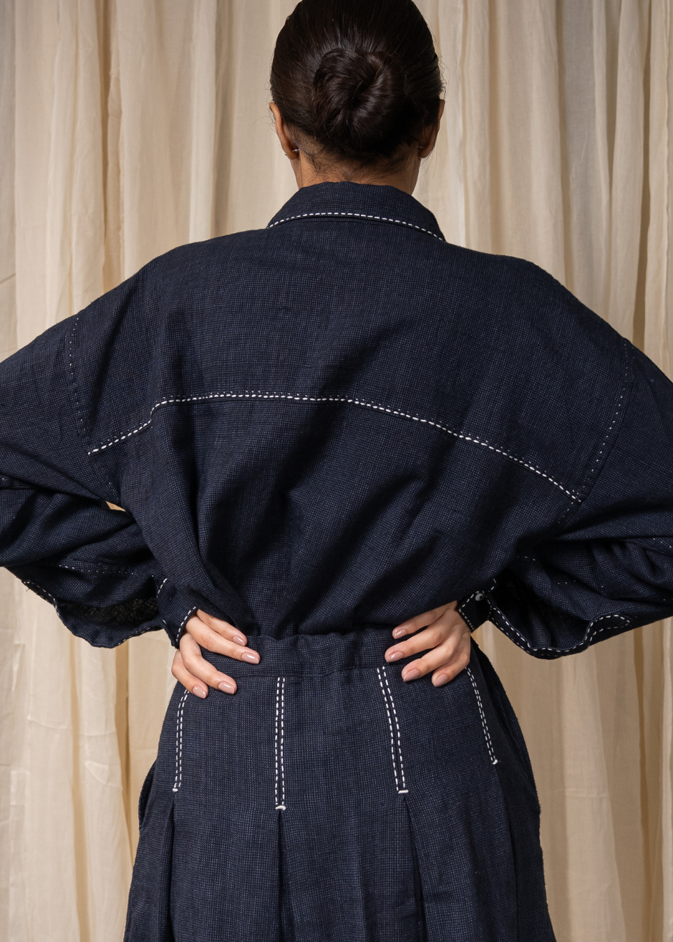 A person wearing a dark indigo denim long-sleeve shirt and matching wide-leg skirt with white contrast stitching, standing against a beige fabric backdrop.