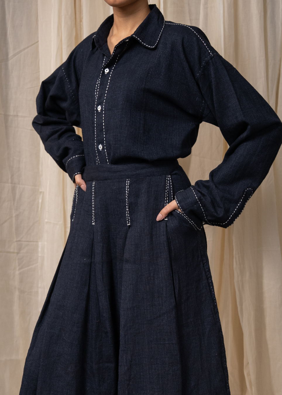 A person wearing a dark indigo denim long-sleeve shirt and matching wide-leg skirt with white contrast stitching, standing against a beige fabric backdrop.