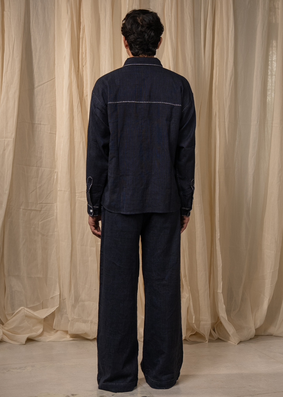 A model viewed from the back wearing a dark indigo denim two-piece set, consisting of a long-sleeve button-up shirt with contrast stitching and matching wide-leg trousers, set against a backdrop of draped beige fabric.
