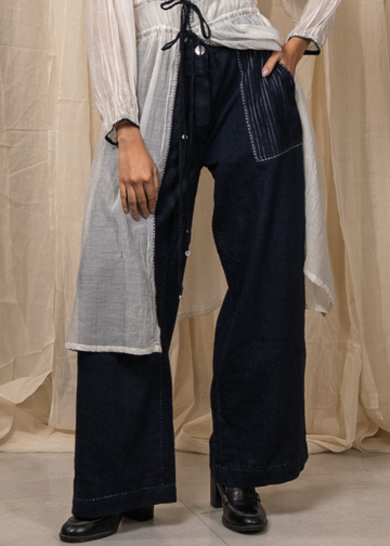 A person wearing dark wide-leg denim pants with contrast stitching and a white sheer long cardigan draped over one side, standing against a beige fabric backdrop.
