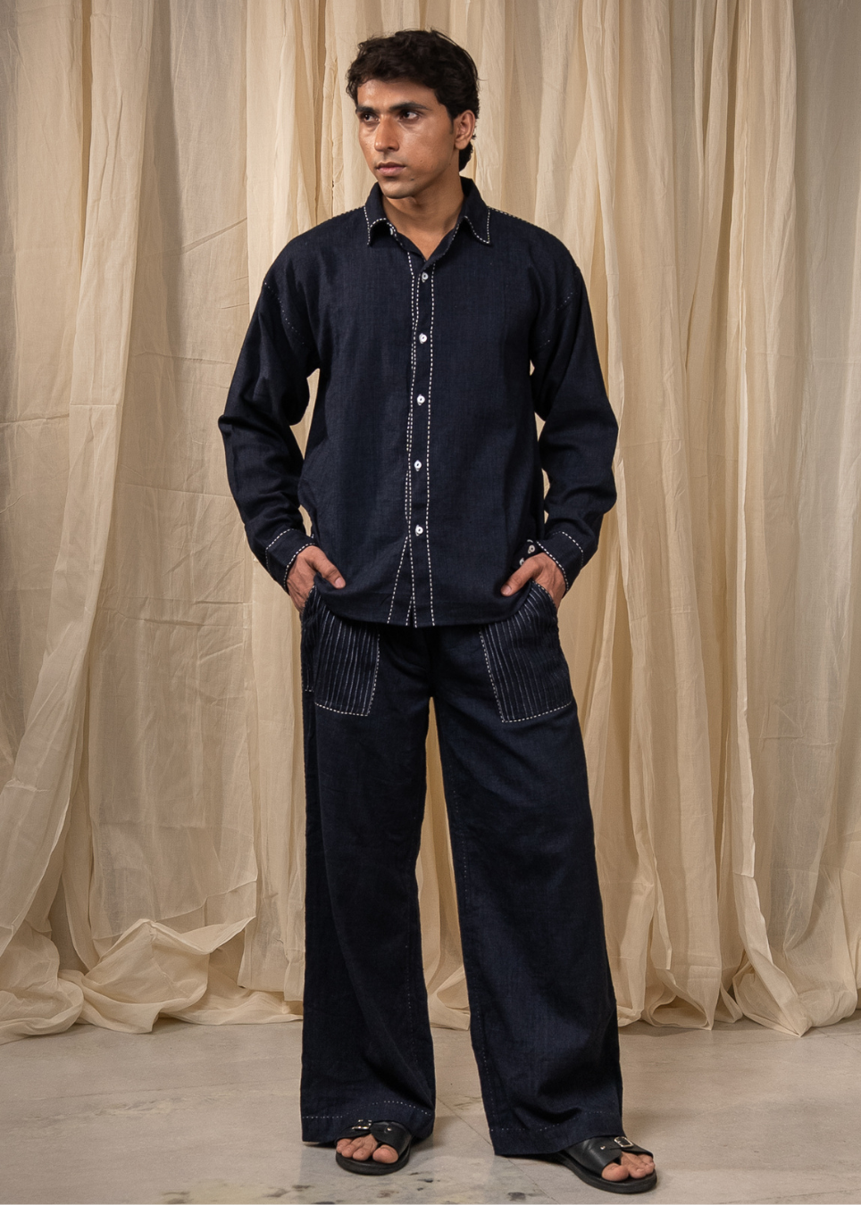 A man wearing a dark indigo denim two-piece set consisting of a long-sleeve button-up shirt and matching wide-leg trousers, standing in front of a draped beige fabric backdrop.