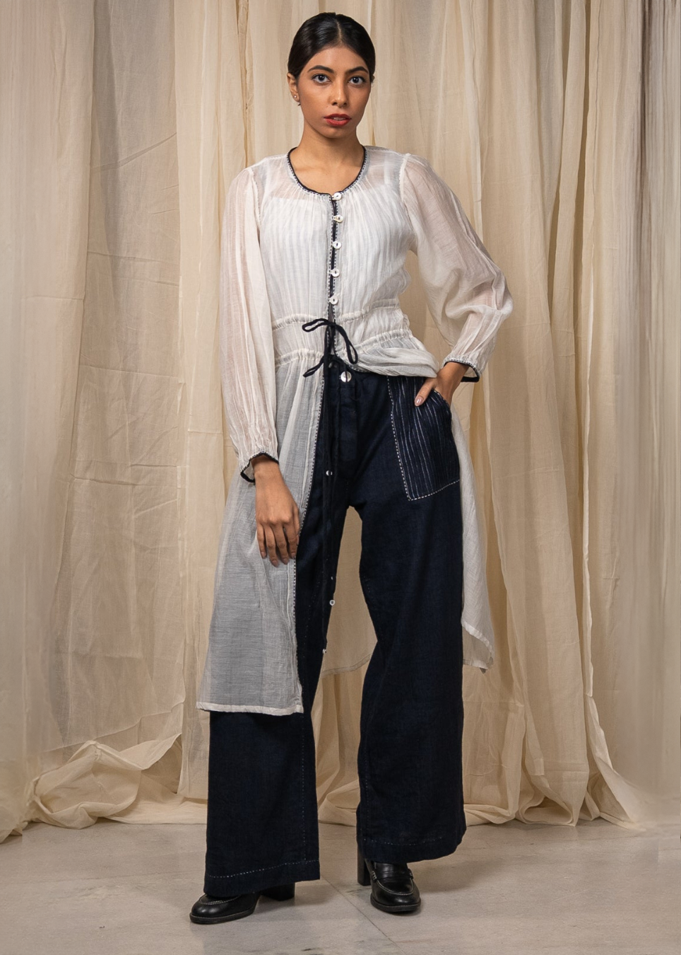 A model stands in front of draped beige fabric, wearing a white long-sleeve top with vertical pleats and a black drawstring, paired with dark wide-leg denim pants.