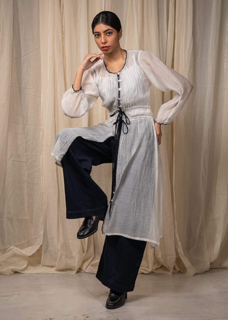 A woman models a white long-sleeve tunic with vertical pleats and a black drawstring waist, paired with dark wide-leg pants, in front of a draped beige fabric backdrop.