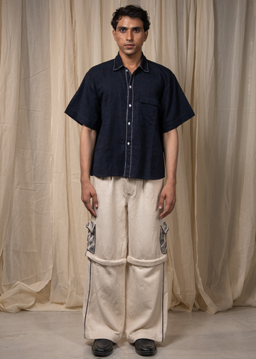 A man stands facing forward wearing a dark navy short-sleeve button-up shirt with white contrast stitching and cream wide-leg pants with dark piping, posed in front of draped beige fabric.