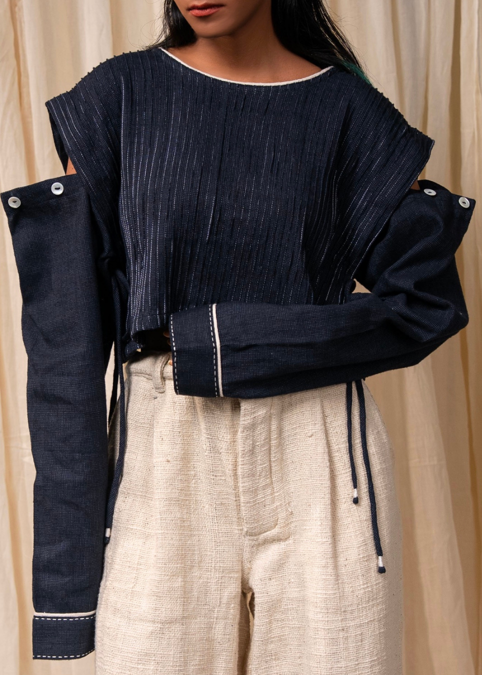A model wearing a dark indigo blue pleated long-sleeve top with contrasting white stitching and oversized buttoned sleeves, paired with light beige linen drawstring pants, set against a neutral beige curtain backdrop.