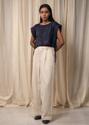 A model stands against a draped beige fabric backdrop wearing a dark navy sleeveless top with side ties and high-waisted, wide-leg cream linen trousers.
