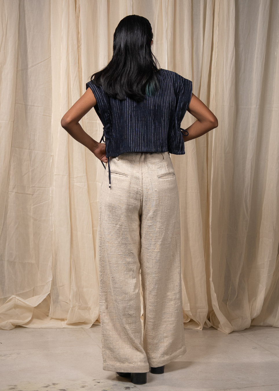 A woman is shown from behind wearing a navy blue sleeveless top and cream wide-leg trousers, standing in front of draped beige curtains.