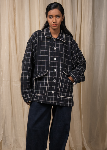 A model stands against a beige curtain backdrop wearing a black and white windowpane plaid button-up jacket with white piping and dark wide-leg jeans.