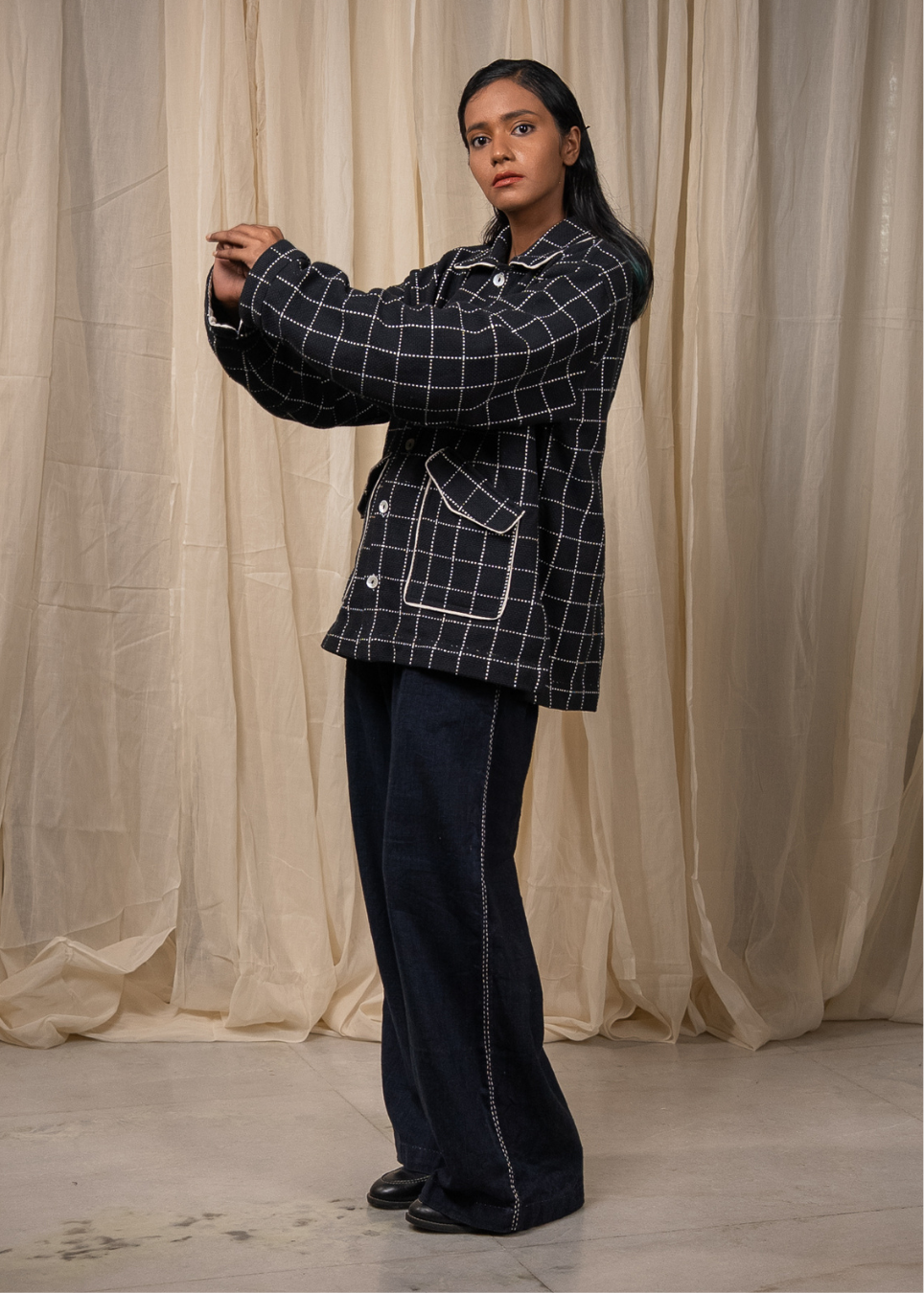 A woman modeling a dark navy blue and white checkered blazer with white piping, paired with dark wide-leg jeans, standing in front of a draped beige fabric backdrop.