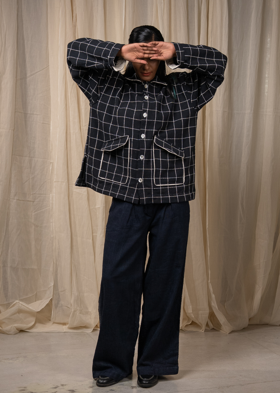 A person wearing a dark navy plaid button-up shirt with white windowpane lines and matching dark wide-leg pants, standing in front of a draped beige curtain backdrop.