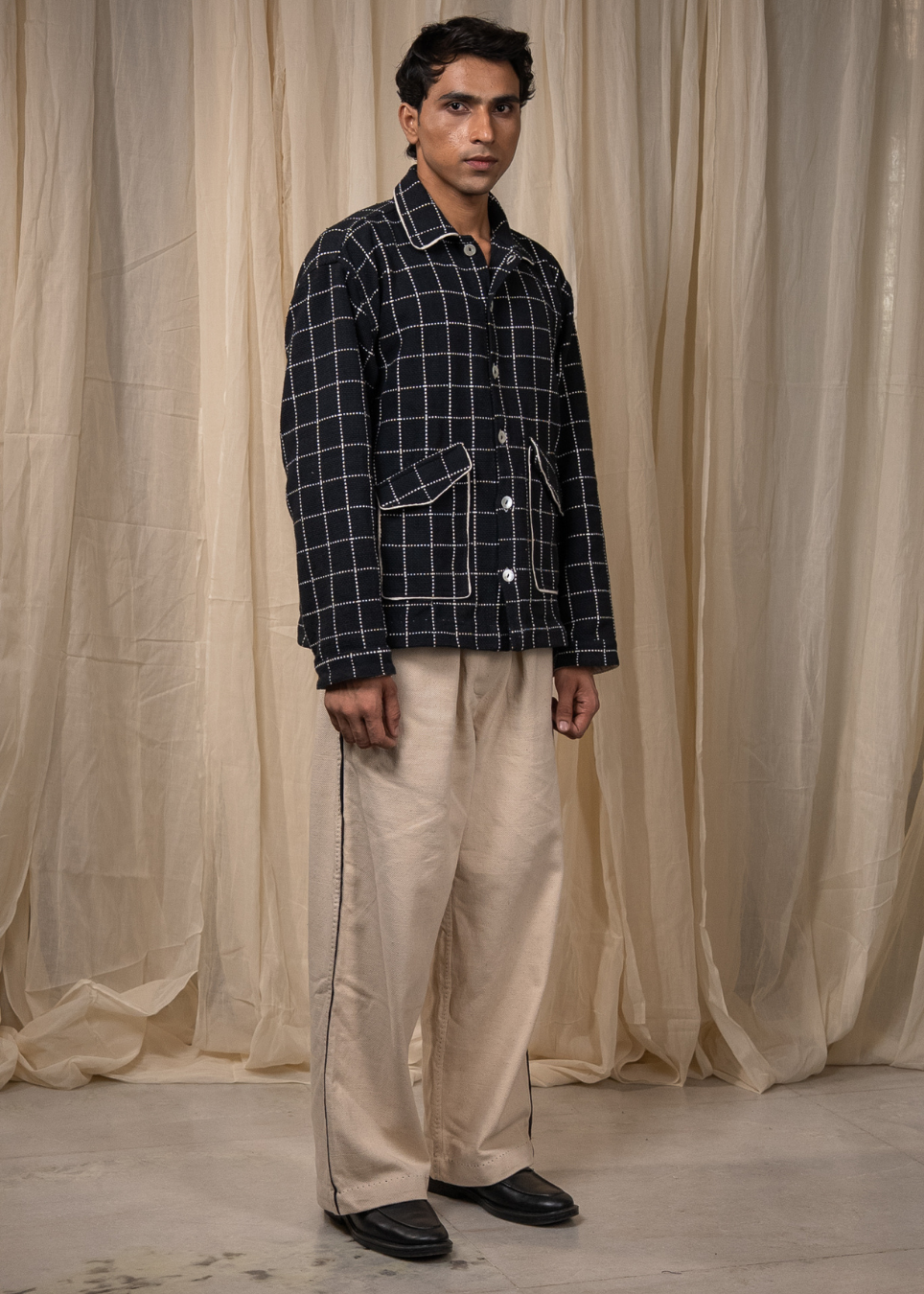 A man stands in front of draped beige fabric, wearing a dark navy and white plaid button-up shirt with beige trousers and black shoes.