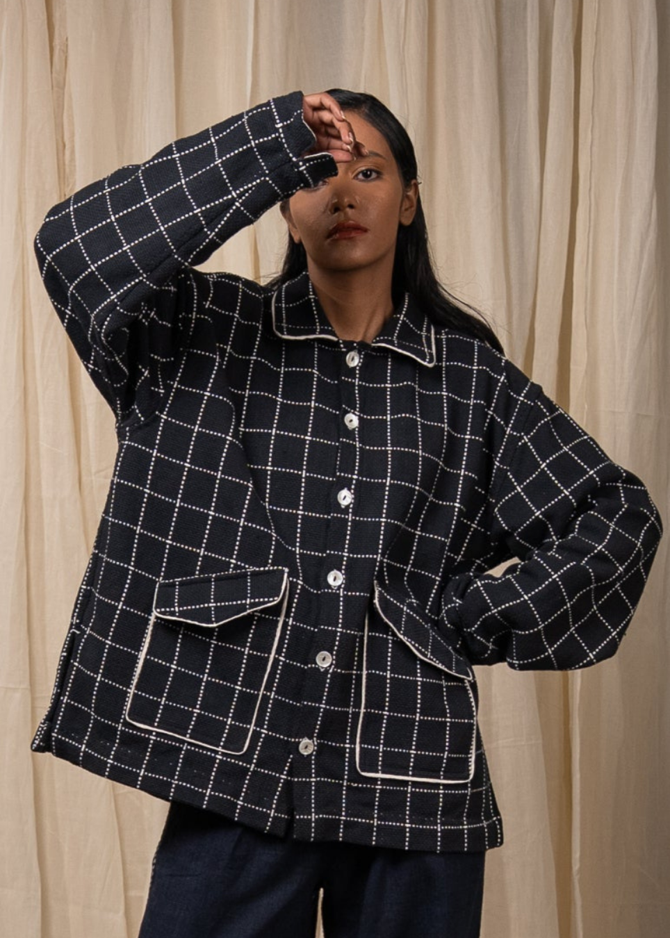 A woman wearing a black quilted jacket with a white windowpane grid pattern and white piping details, posing in front of a beige curtain backdrop.