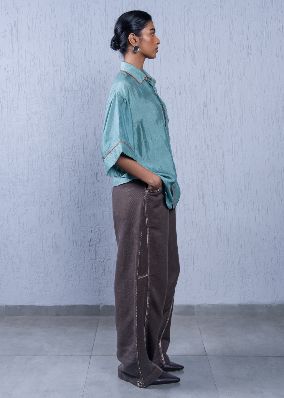 A woman stands in profile wearing a light teal short-sleeve button-up shirt with contrast stitching and dark brown wide-leg trousers with visible seam detailing, set against a textured light gray wall and tiled floor.