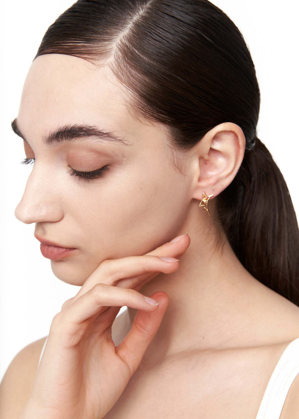 A close-up of a woman with dark hair in a ponytail, wearing a delicate gold crescent moon earring on her earlobe, set against a plain white background.