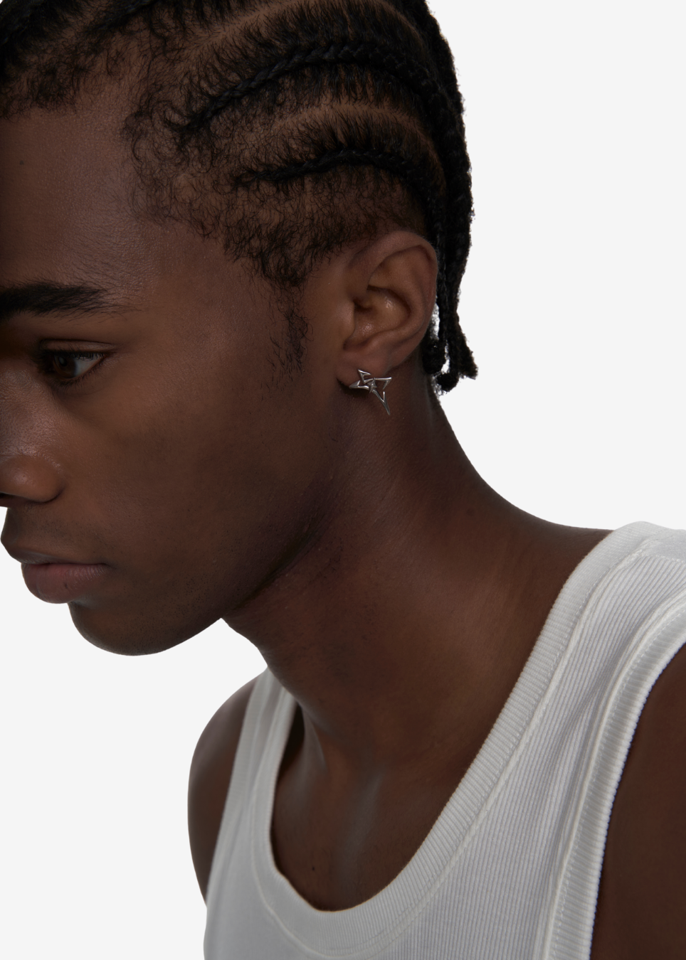 A close-up side profile of a person with braided hair wearing a small silver earring with a dangling star-shaped element, paired with a white ribbed tank top against a plain light background.
