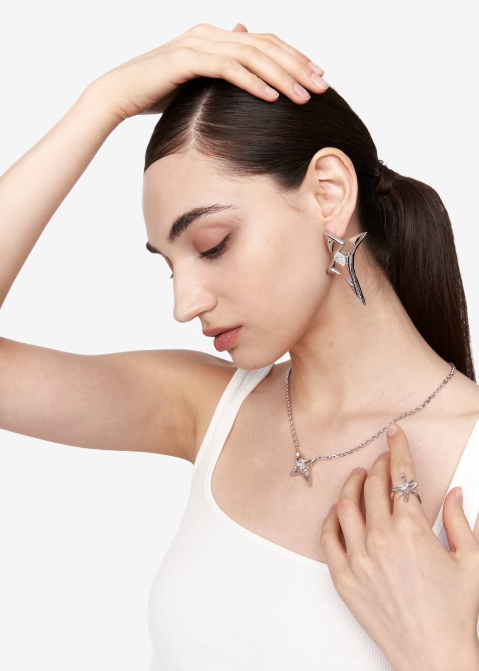 A woman with dark hair in a ponytail models a silver-toned jewelry set featuring a matching necklace and earring with star-shaped crystal details, set against a clean white background.
