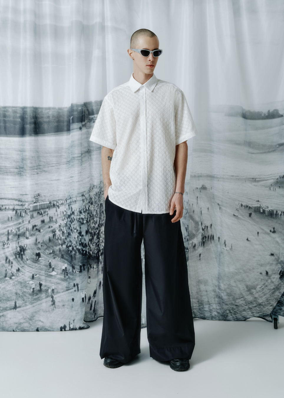 A model stands wearing a white short-sleeve button-up shirt with a subtle floral pattern, paired with wide-leg black trousers and black sunglasses, set against a large black and white landscape photograph backdrop.