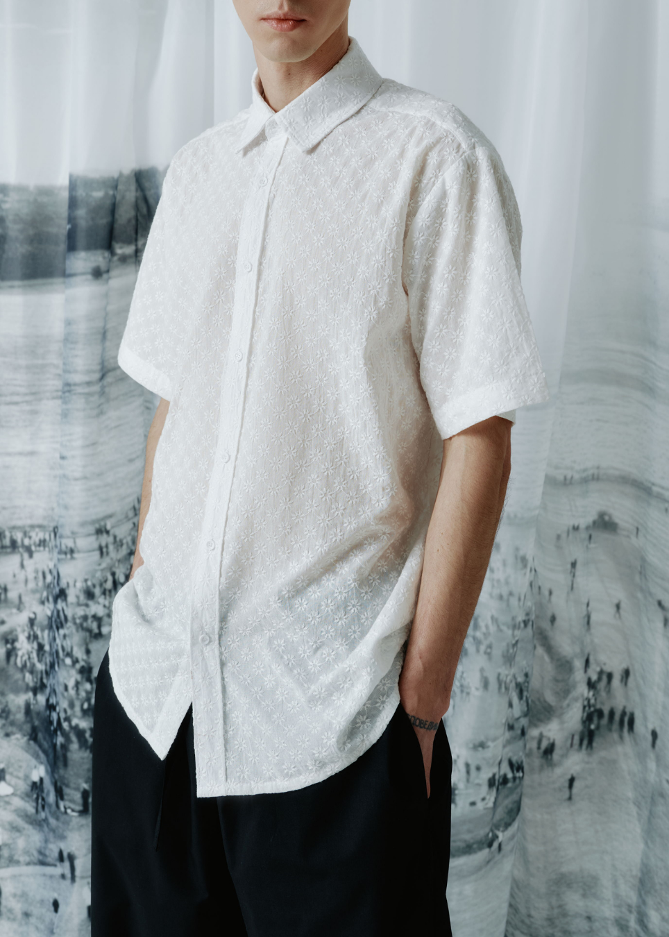 A man wearing a white short-sleeve button-up shirt with a subtle textured pattern, paired with black trousers, standing in front of a gray and white scenic backdrop.