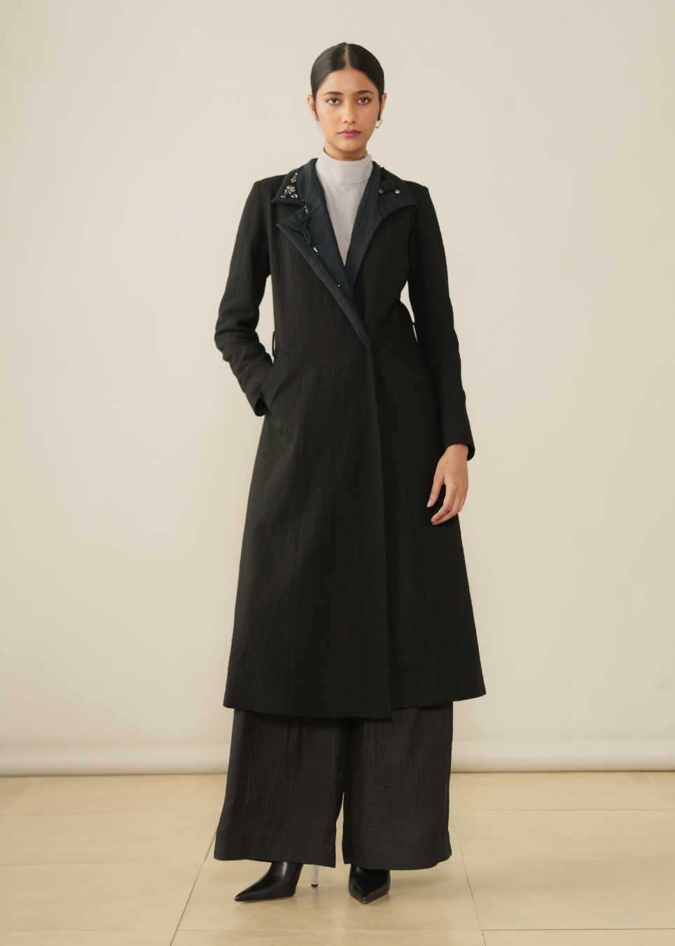 A model stands in a studio wearing a long black coat over a white mock neck top and wide-leg black trousers.