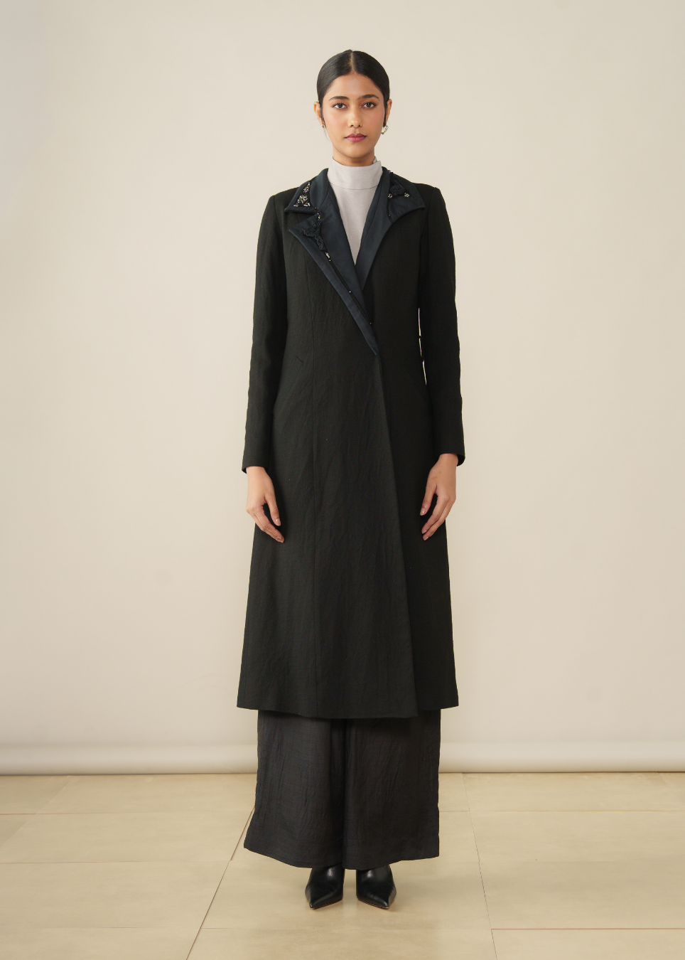 A model stands in a studio wearing a long black coat over a white turtleneck and wide-leg black trousers. The coat features a notched collar with dark blue fabric lining and decorative silver-toned embellishments.
