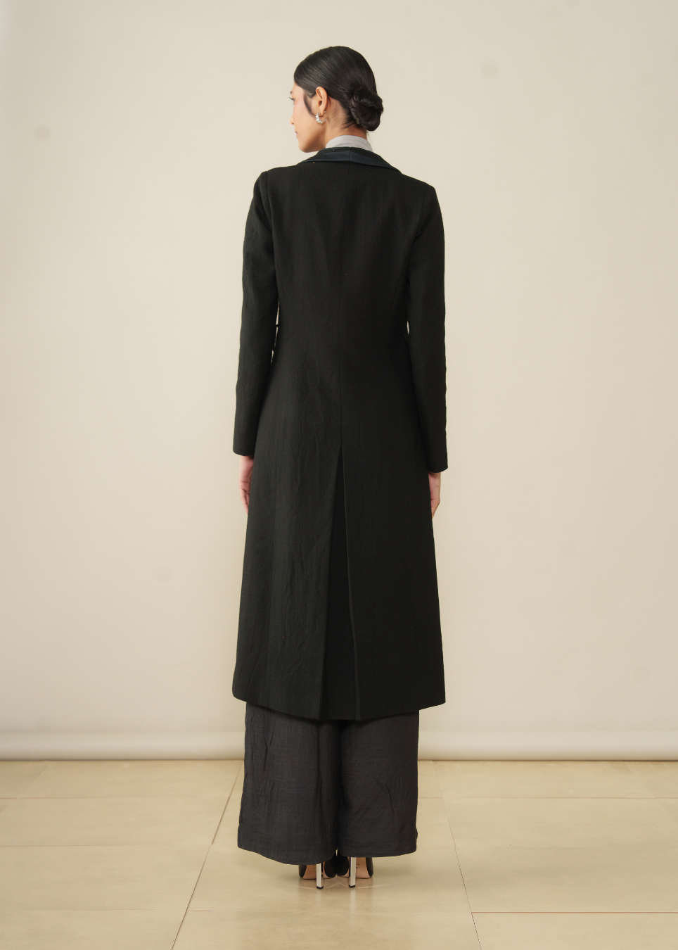 A model viewed from the back wearing a long black coat over dark wide-leg pants, standing against a plain light beige wall and floor.