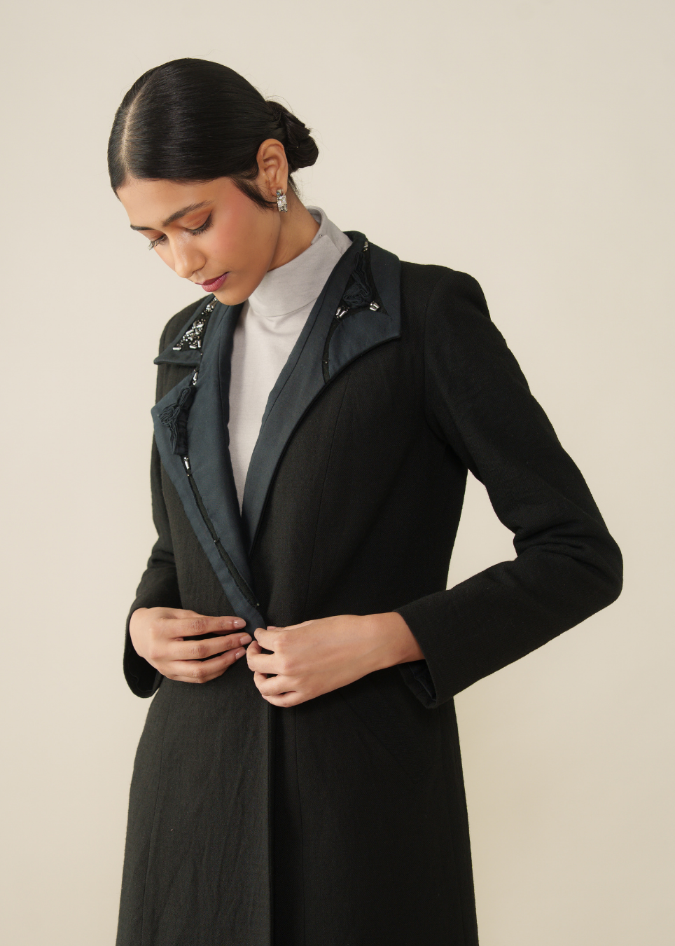 A woman wearing a black long coat with a dark gray inner collar and zipper detail, layered over a white mock neck top, posing against a solid light beige background.