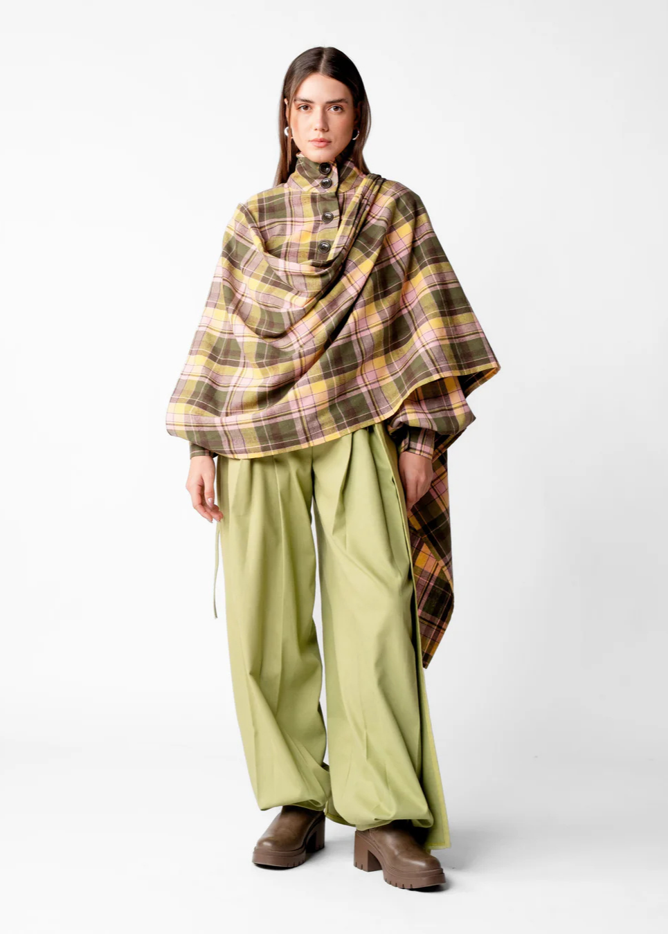 A model stands against a light gray studio background wearing a large plaid shawl draped over her shoulders, olive green wide-leg pants, and brown block-heel boots.
