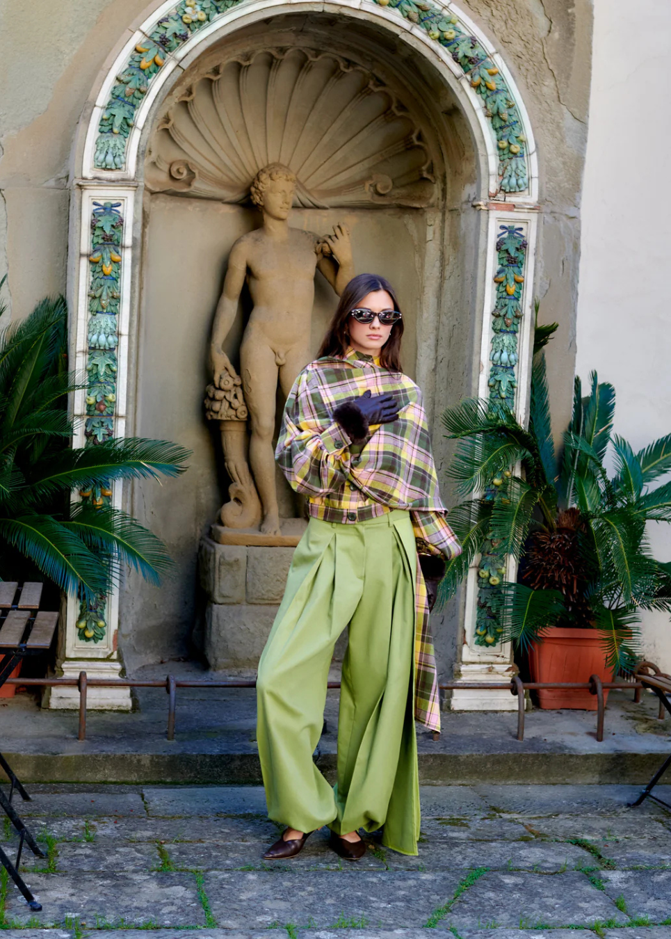 A woman posing in front of a stone archway with a classical statue, wearing a yellow and brown plaid shirt, bright green wide-leg trousers, and dark sunglasses.