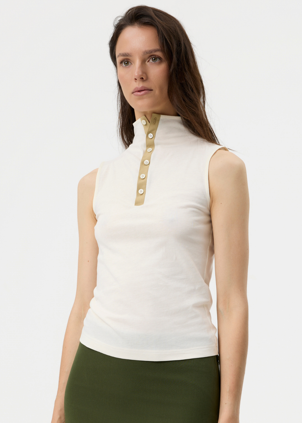 A woman modeling a sleeveless white mock neck top with a beige placket and decorative buttons, paired with an olive green skirt, against a plain white studio background.