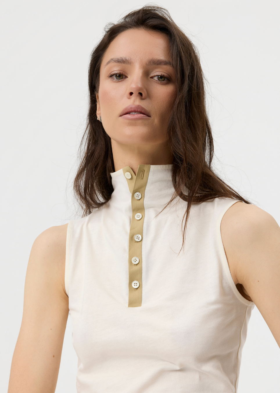 A woman wearing a sleeveless cream-colored top with a high mock neck and a vertical beige button placket, photographed against a light gray studio background.