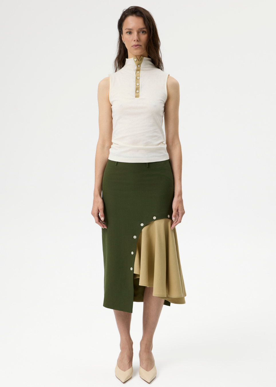 A model stands against a plain white studio background wearing a sleeveless white mock-neck top with gold trim and an asymmetrical olive green skirt with a gold panel and white buttons.