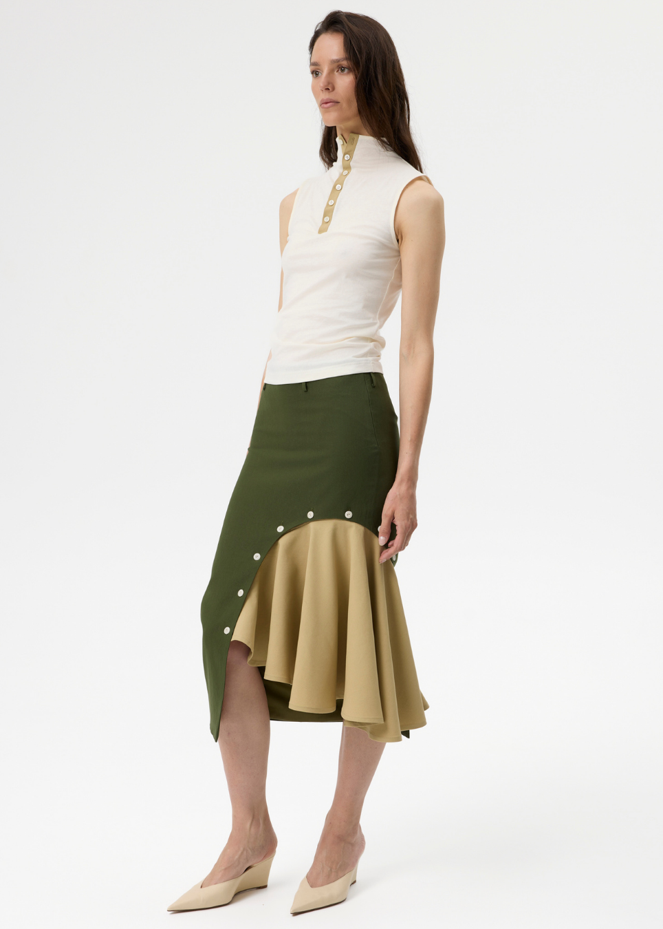 A model wearing a sleeveless white mock-neck top with gold trim and a two-tone midi skirt in olive green and beige with decorative buttons, paired with beige wedge heels, against a plain white studio background.