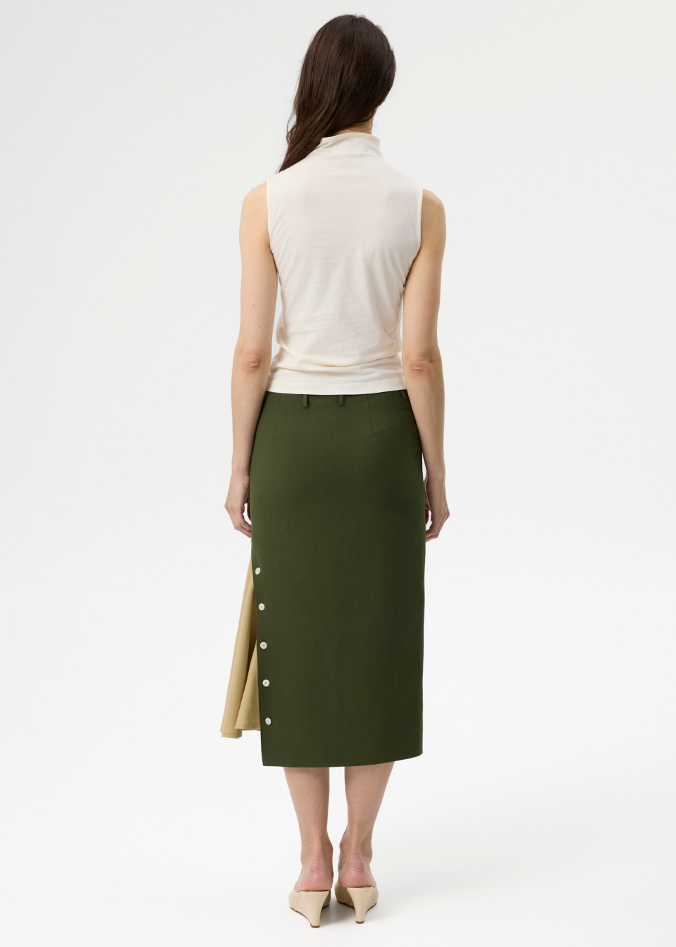 A woman shown from the back wearing a sleeveless cream mock-neck top and an olive green midi skirt with a beige side panel and decorative buttons, against a plain white studio background.